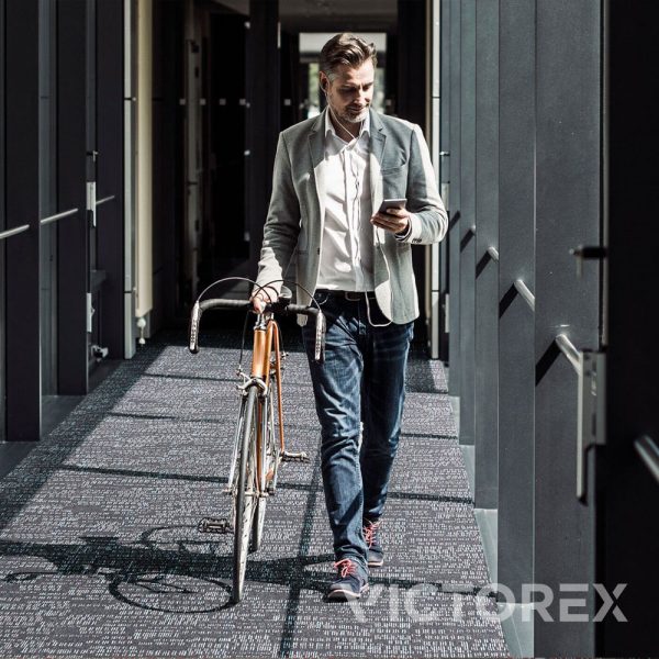 Interface Works Atom carpet tile in Cobalt colour, with a guy walking while holding phone and bicycle; Brick layout.